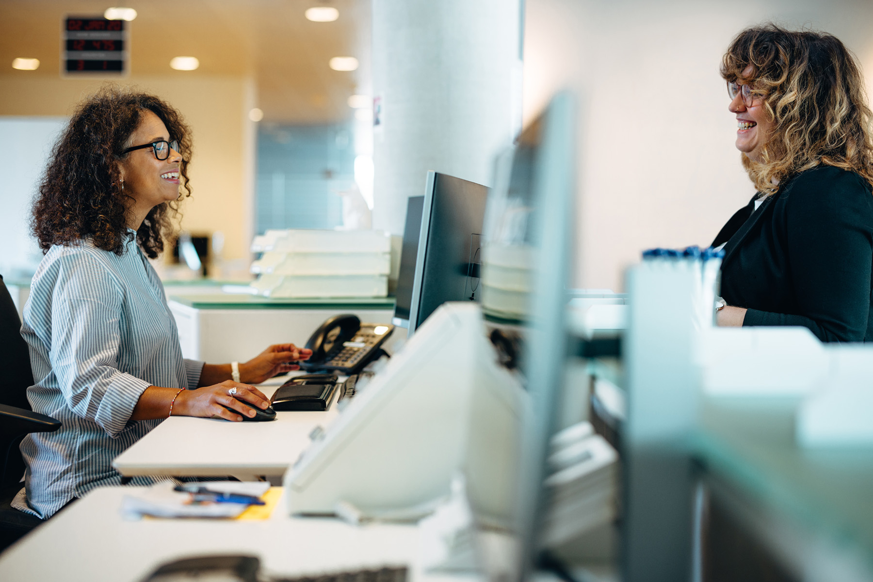 Zwei Frauen stehen an einem Schalter in einem modernen Büro. Eine der Frauen sitzt hinter dem Schalter, während die andere, die vor dem Schalter steht, freundlich mit ihr spricht. Beide tragen Brillen und lächeln.